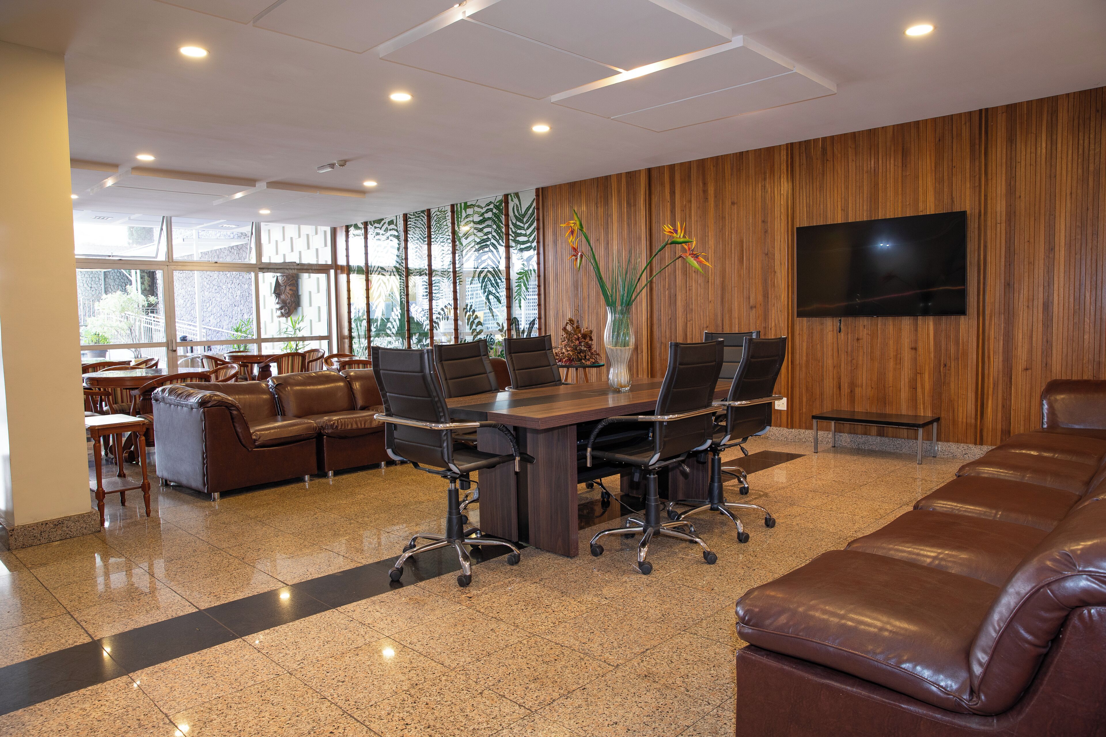 A conference room with a wooden panel wall, a large flat-screen TV, a brown leather sofa, and a dining area with wooden furniture.