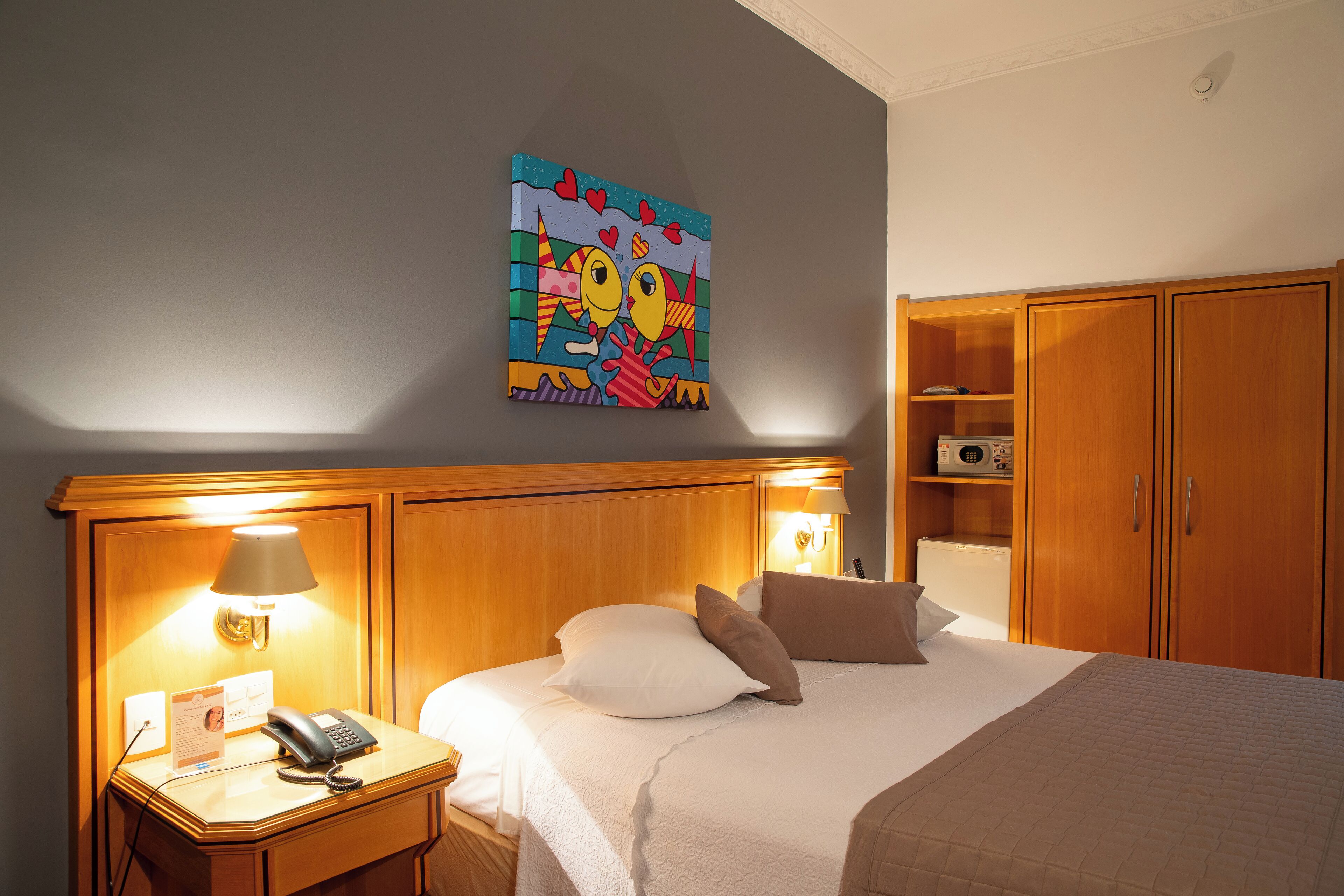 Una habitación de hotel con una cama, lámparas de noche, un escritorio con teléfono y un cuadro en la pared.