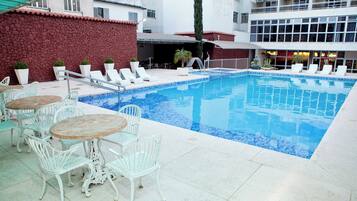 A swimming pool with lounge chairs and tables, surrounded by a stone wall and a building with large windows.