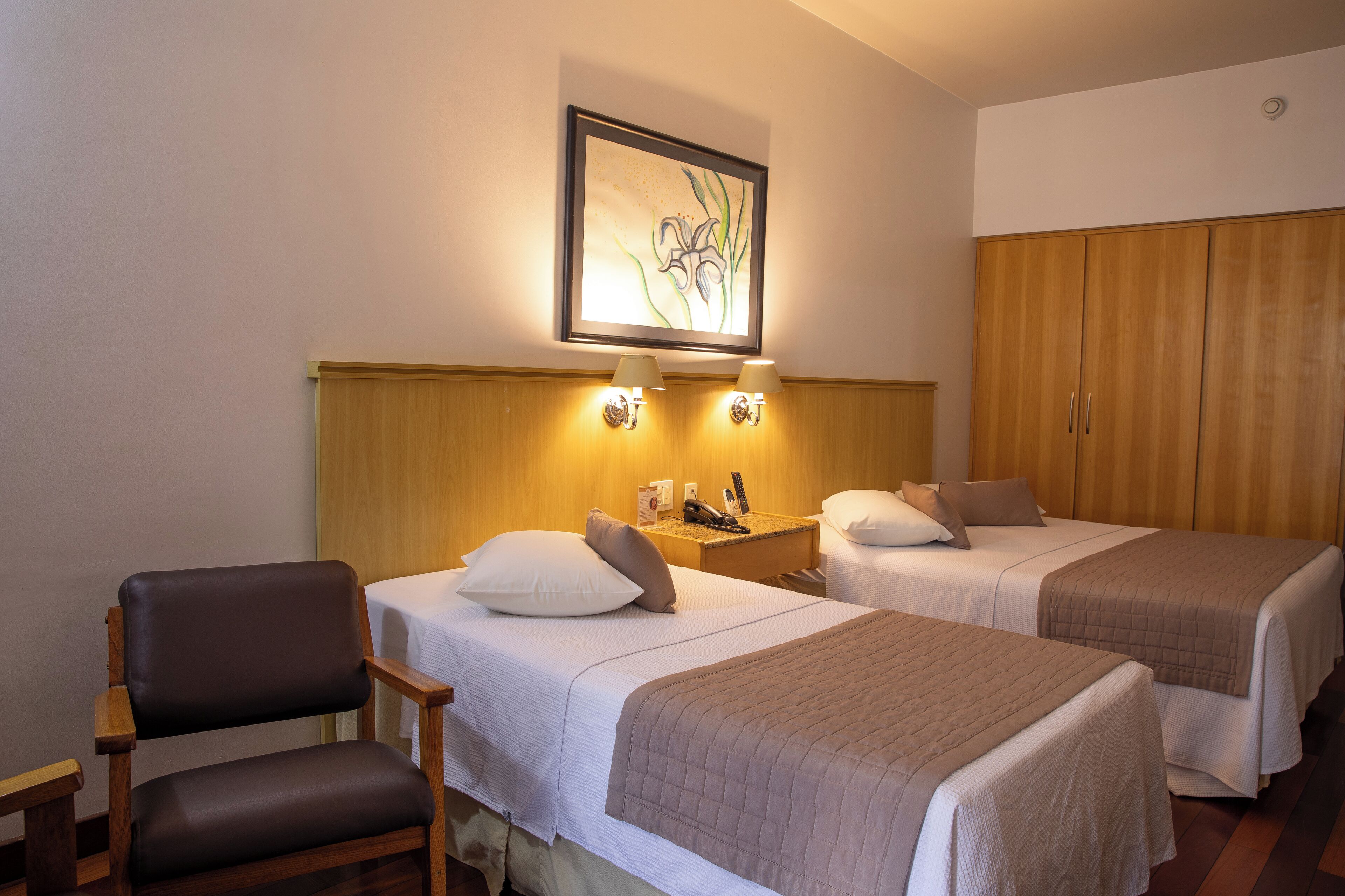 A hotel room with two beds, a chair, a nightstand, and a wardrobe.