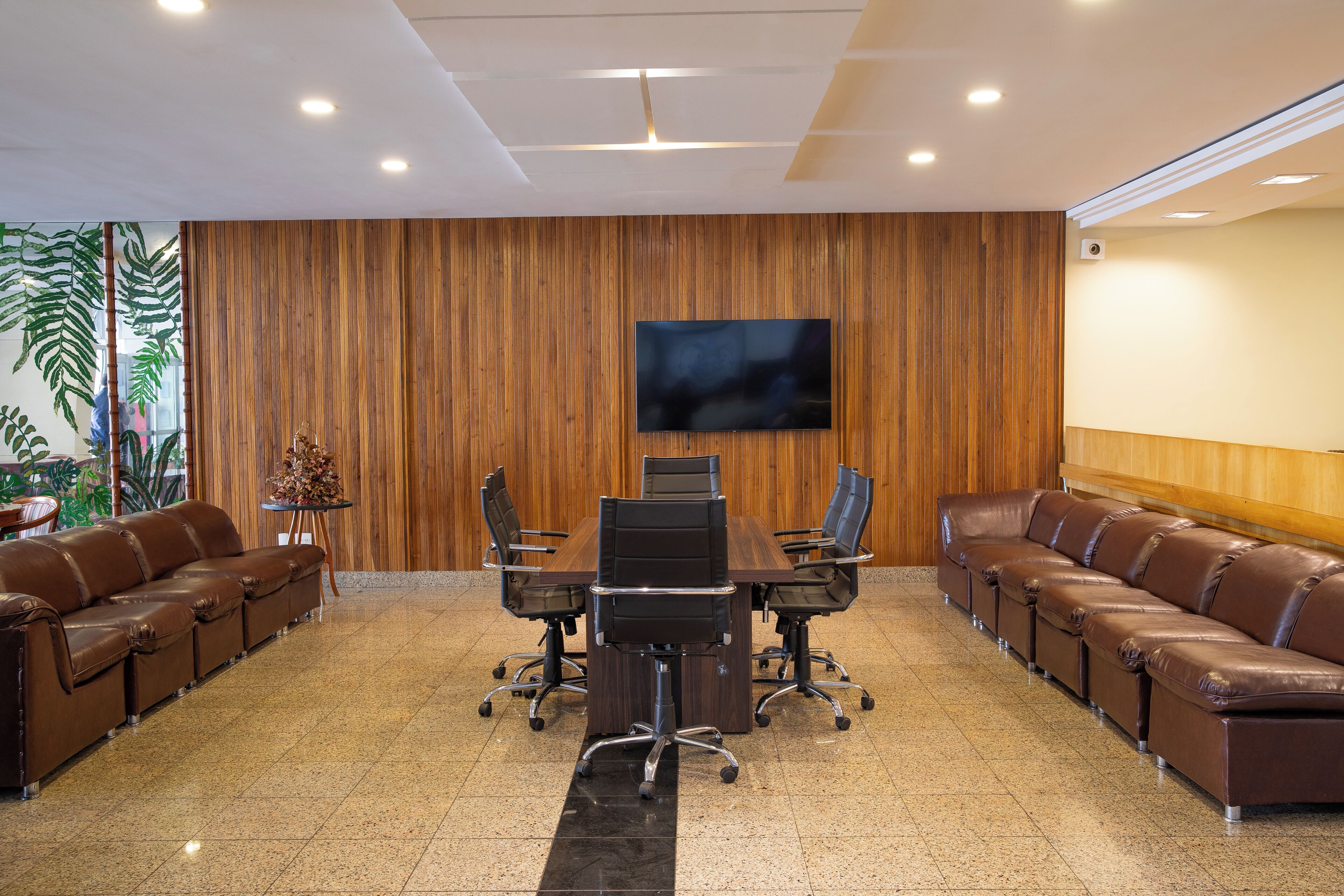 Una sala de conferencias con pared de paneles de madera, un televisor de pantalla plana grande, un sofá de cuero marrón largo y una mesa redonda con sillas.