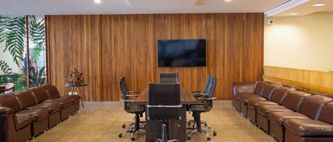 Una sala de conferencias con pared de paneles de madera, un televisor de pantalla plana grande, un sofá de cuero marrón largo y una mesa redonda con sillas.
