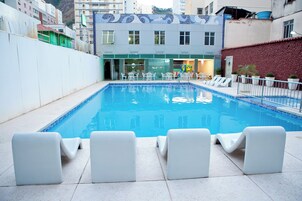Piscina com cadeiras de descanso brancas e um edifício com fachada revestida ao fundo.