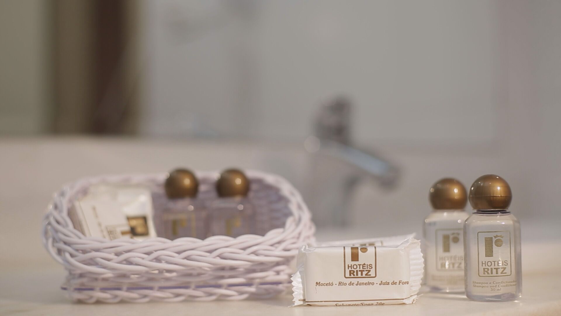 A hotel minibar setup with a wicker basket containing bottles, a minibar box labeled 'HOTÉIS RITZ,' and two bottles of 'HOTÉIS RITZ' shampoo.