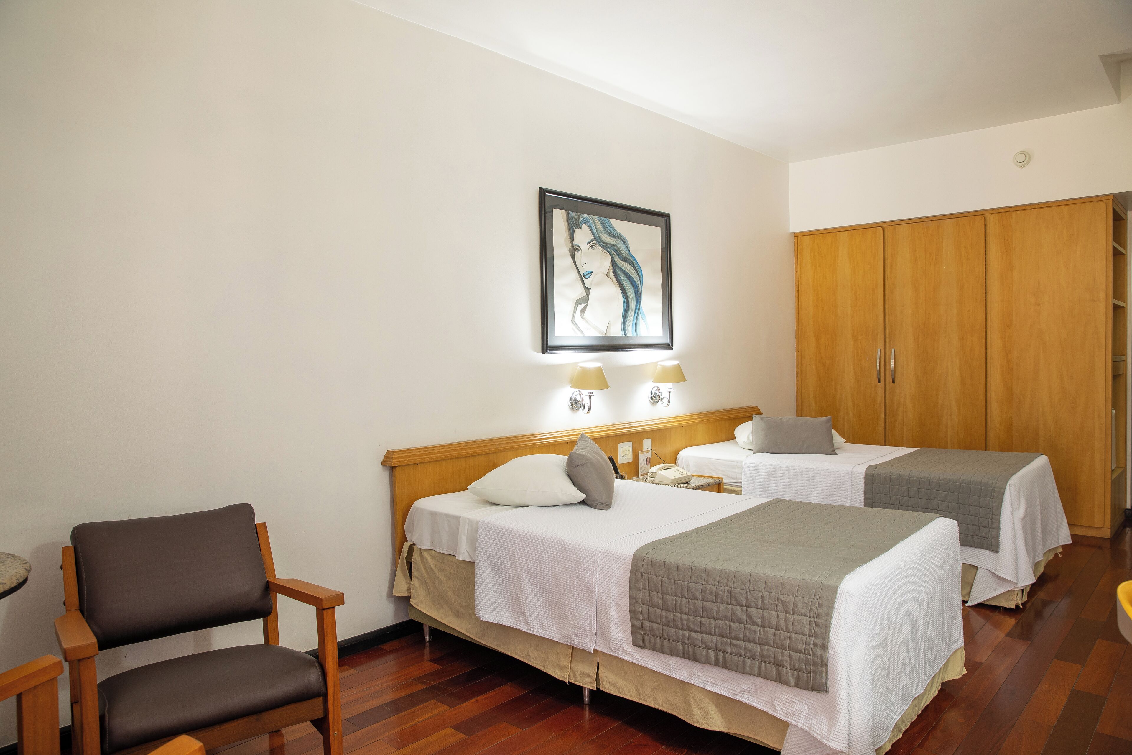 A hotel room with two beds, a wooden wardrobe, a chair, and a painting on the wall.