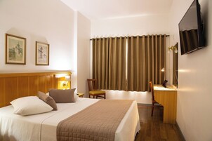 Quarto de hotel com cama, escrivaninha, cadeira, televisão e cortinas.