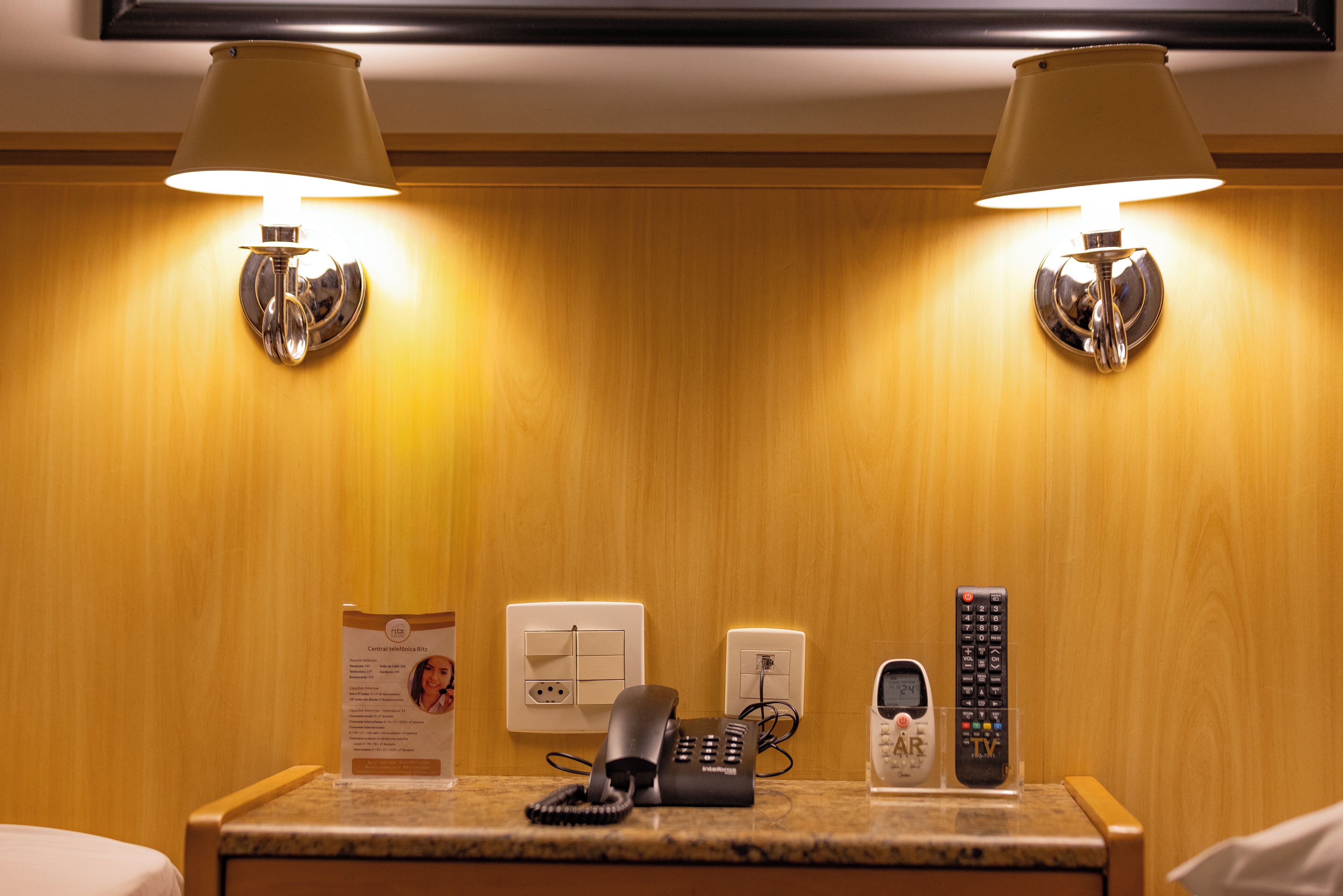 A hotel room with a wooden wall, a bedside table with a phone, a remote control, and a lamp.