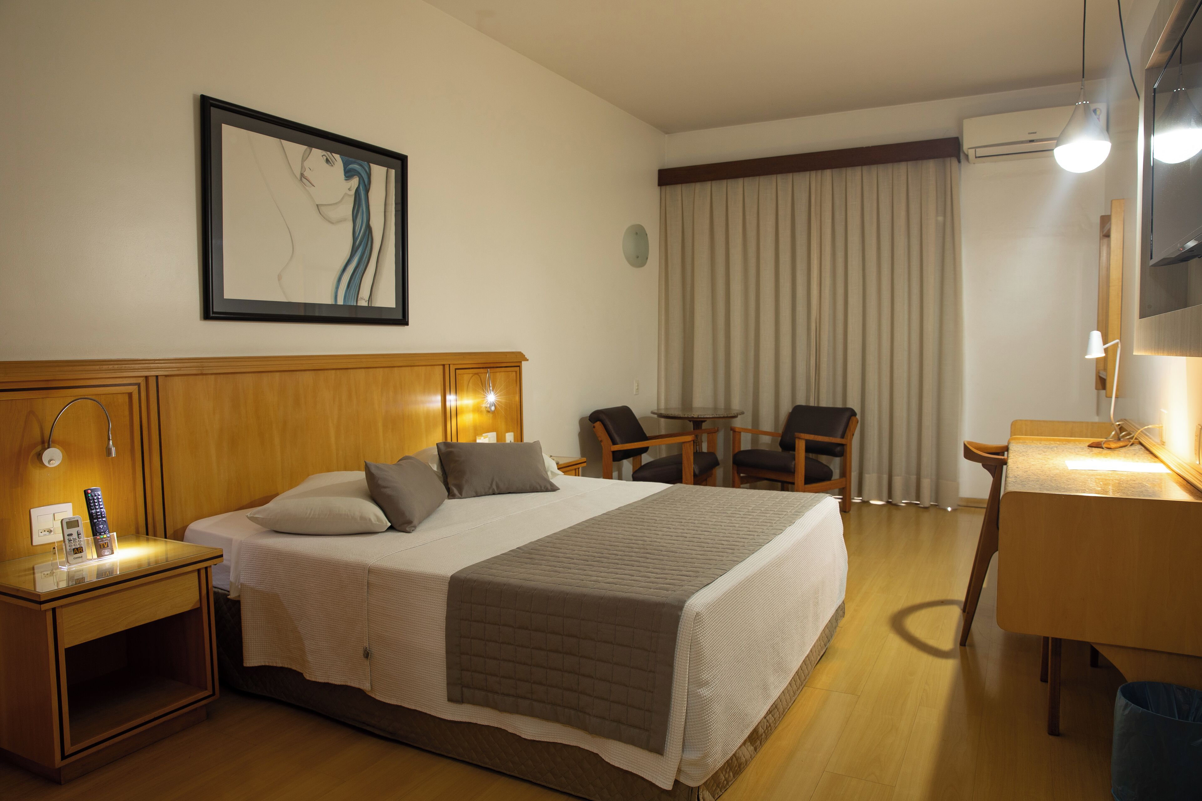 Une chambre d’hôtel avec un lit, une table de chevet, une lampe, un bureau, une chaise et un tableau au mur.
