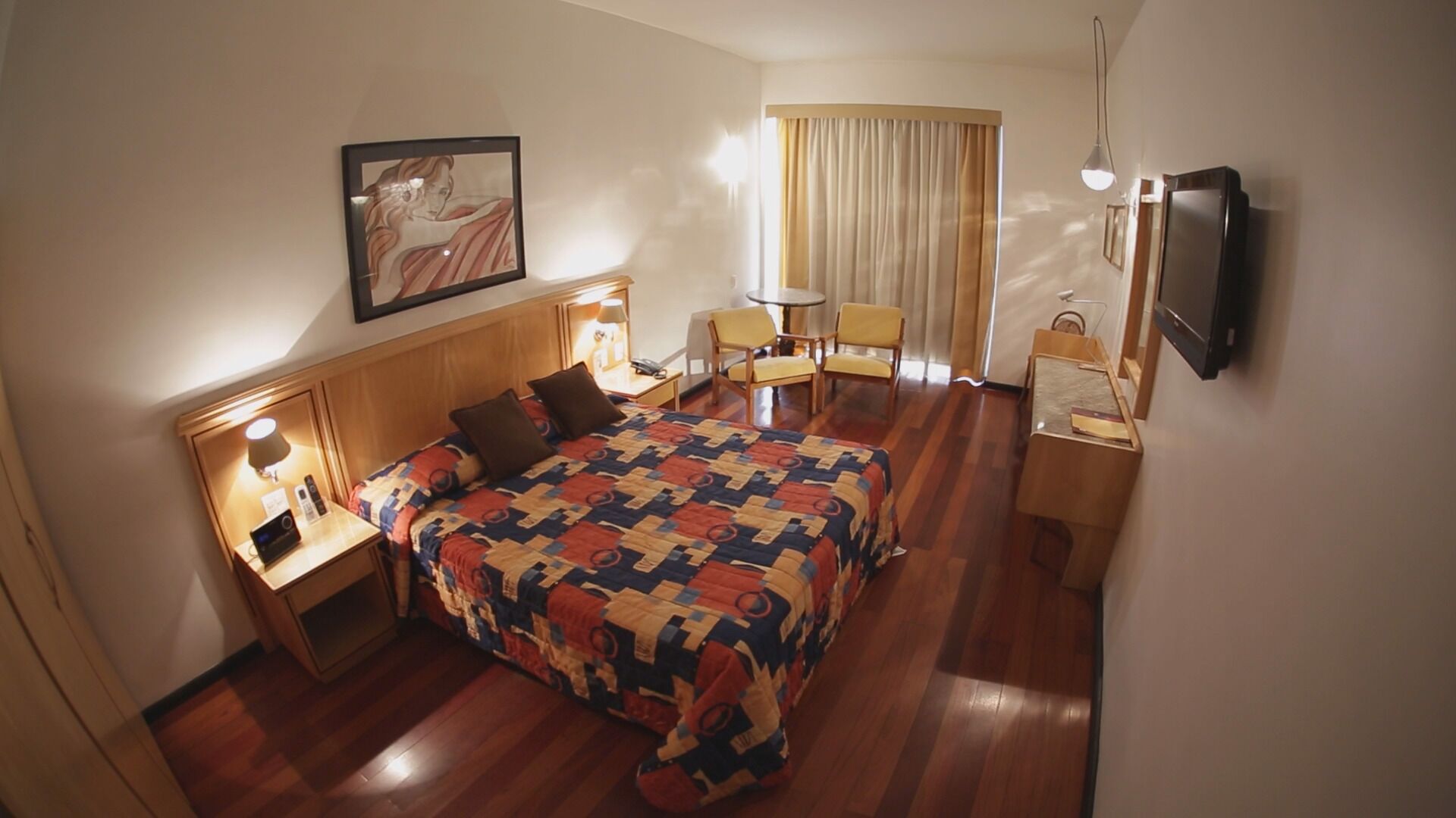 Habitación de hotel con una cama grande, un escritorio con lámpara, una silla, televisión y un cuadro en la pared.