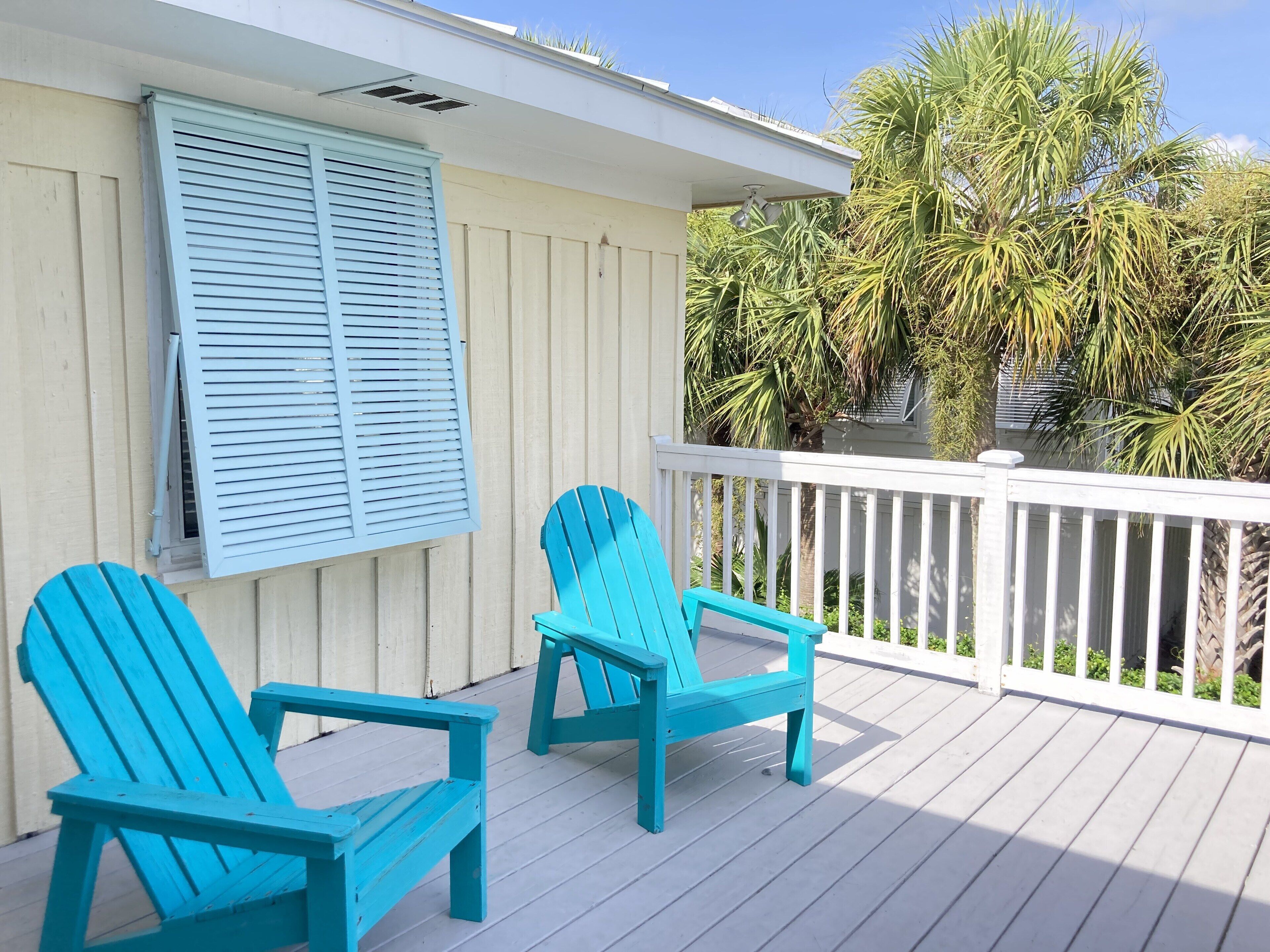 A deck with two turquoise Adirondack chairs, a white railing, and a view of palm trees.