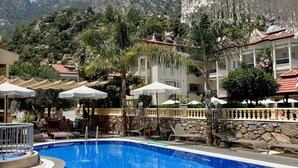 Una piscina con sillas de descanso y sombrillas, rodeada de edificios y montañas.