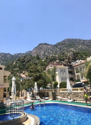 A swimming pool with lounge chairs and umbrellas, surrounded by buildings and a mountainous backdrop. - Gozdem Apart 2 (Marmaris)