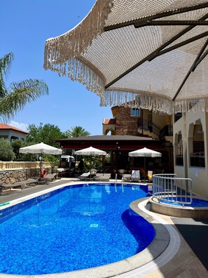 A swimming pool with lounge chairs and umbrellas, surrounded by a stone wall and a building with a tiled roof. - Gozdem Apart 2 (Marmaris)