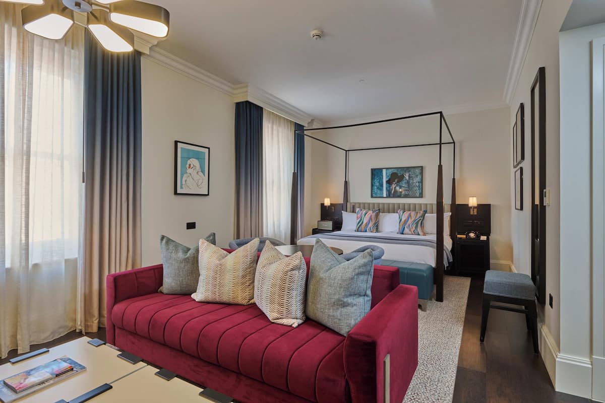 Une chambre d’hôtel moderne avec un canapé rouge, un lit à baldaquin, une table de chevet et un petit banc.