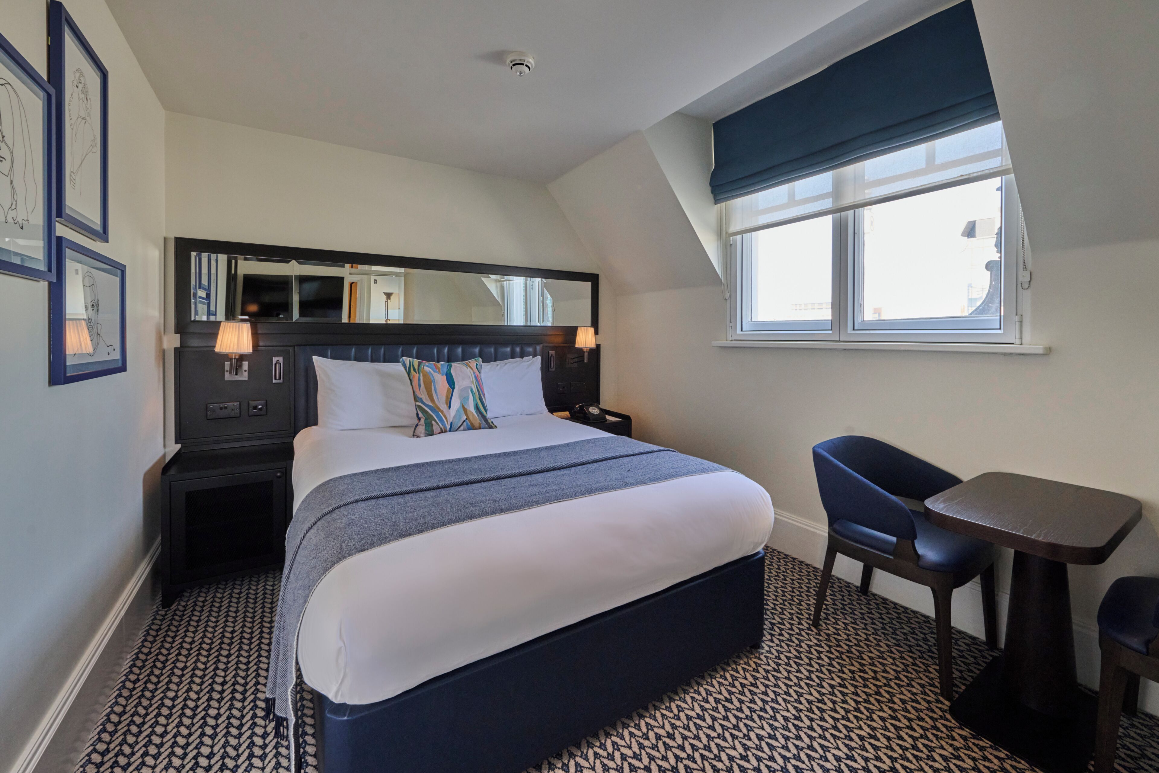 Habitación de hotel con una cama grande, un escritorio, una silla, una ventana con vistas y un cabecero con espejo y lámparas.