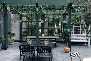 An outdoor patio with a table and chairs, surrounded by greenery and a white bench.
