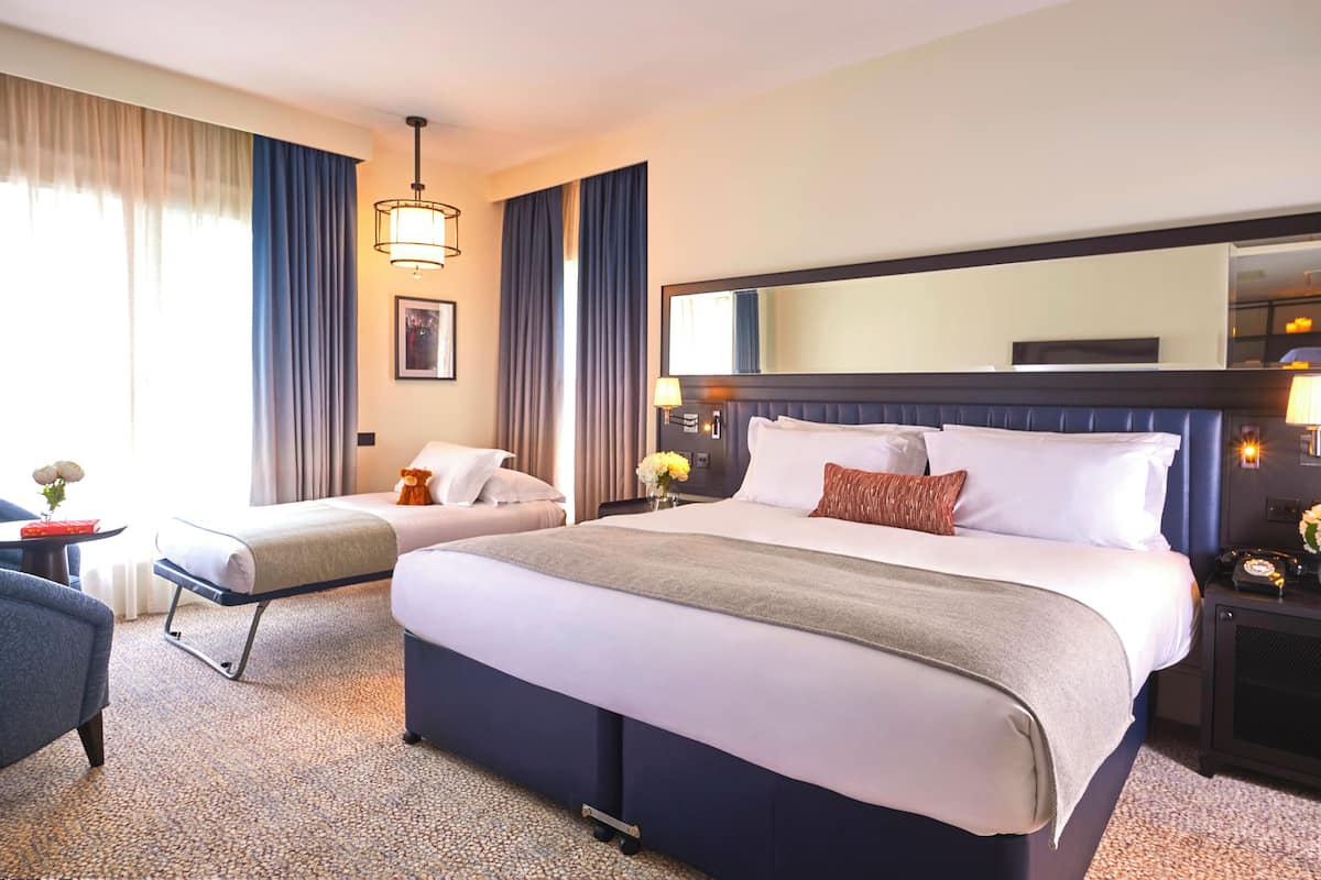 Une chambre d’hôtel avec un grand lit, une table de chevet, une chaise, un bureau et une grande fenêtre avec des rideaux.