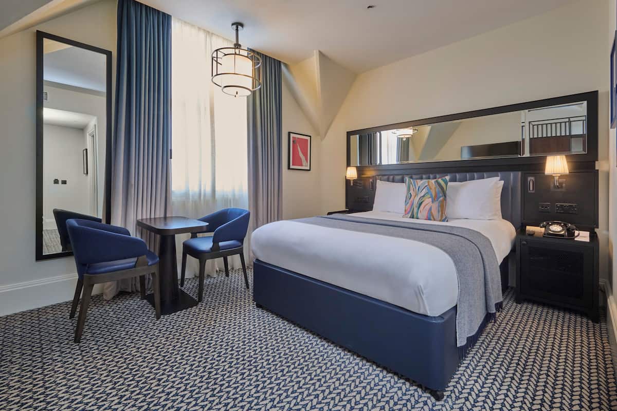 Une chambre d’hôtel avec un grand lit, deux chaises, une petite table, un miroir et une table de chevet avec une lampe.