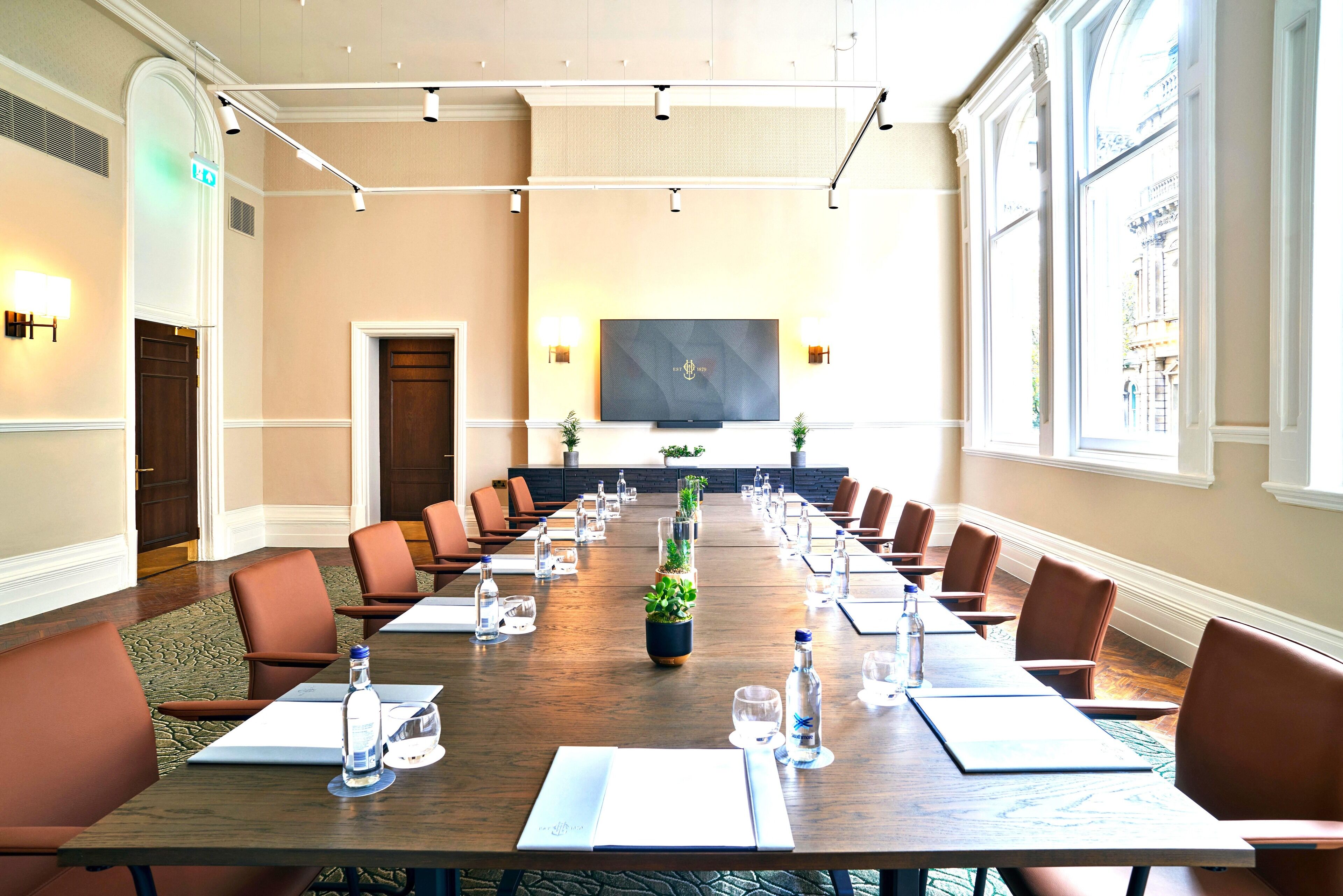 A conference room with a long table, brown leather chairs, water bottles, and a large screen at the front.