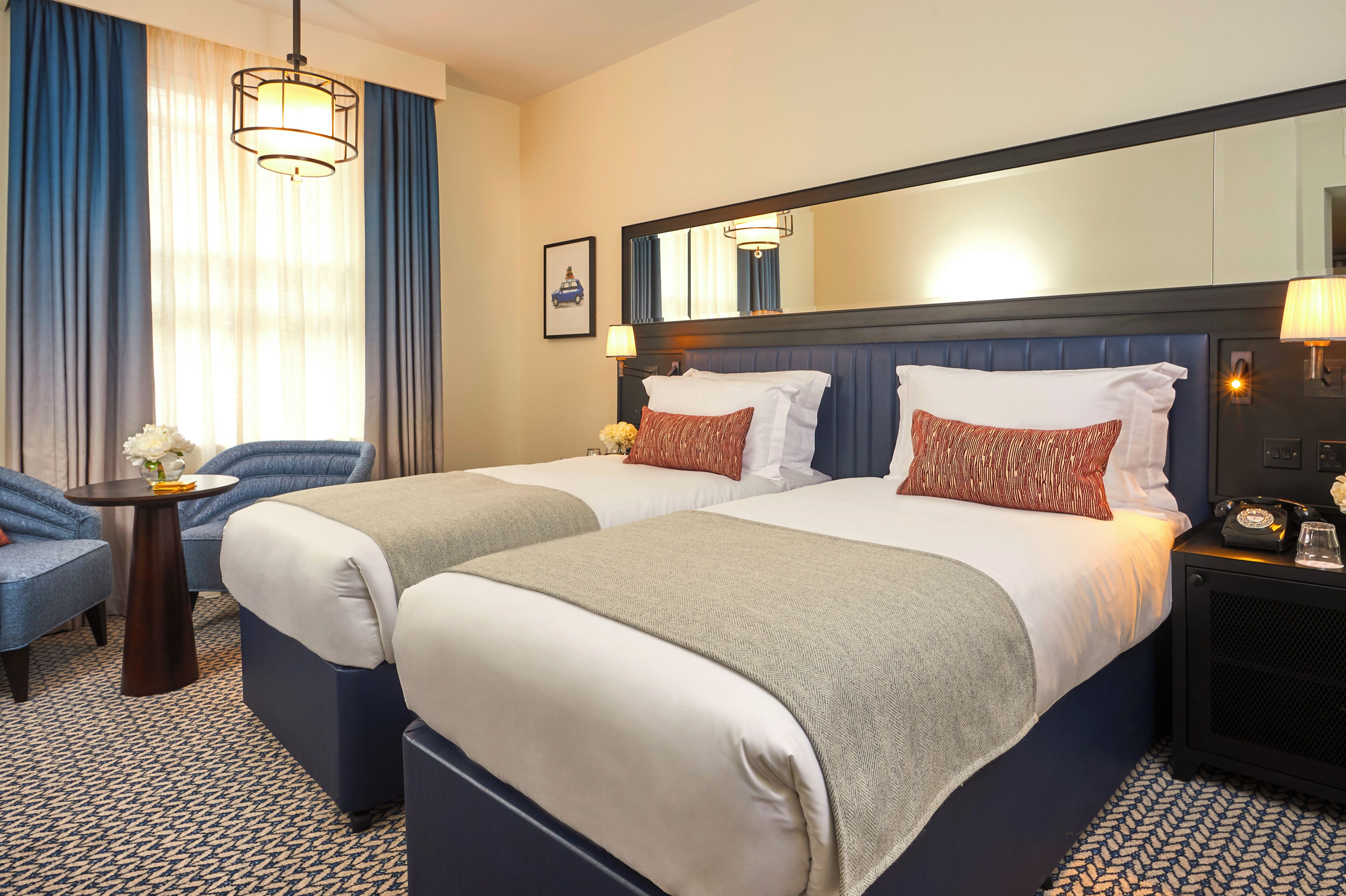 A hotel room with two beds, a desk, a chair, a lamp, and a mirror.