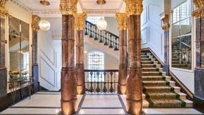 A grand staircase with ornate columns and a chandelier, leading to a second-floor landing with a glass railing.