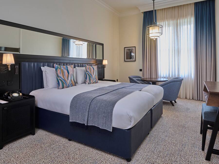 A hotel room with a large bed, a desk, a chair, a lamp, a telephone, and a mirror.