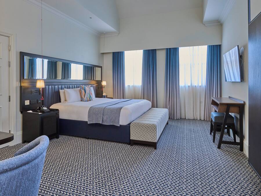 A hotel room with a bed, bedside table, lamp, chair, desk, and large windows with curtains.