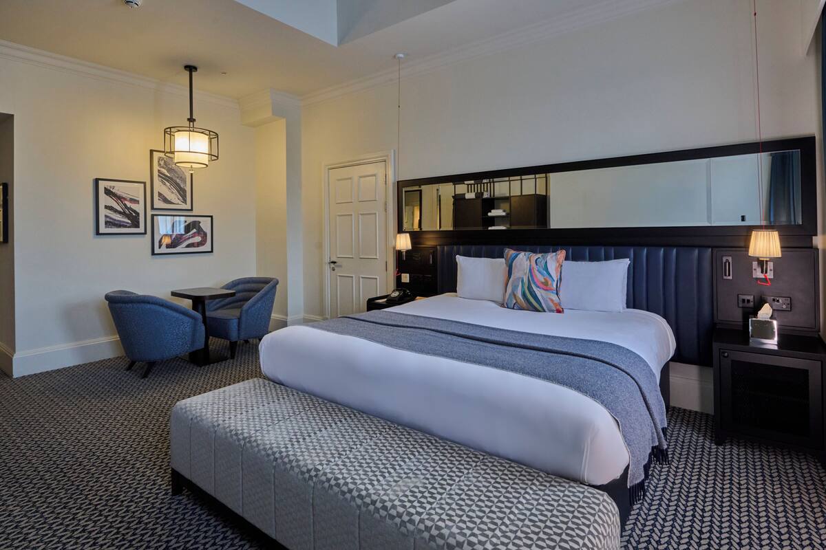Une chambre d’hôtel avec un grand lit, un bureau, deux chaises, une table de chevet, un miroir et des œuvres d’art accrochées au mur.