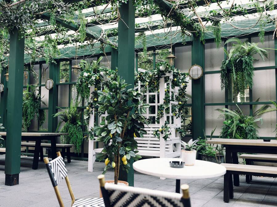 An indoor garden with wooden benches and tables, surrounded by green plants and a white fence.