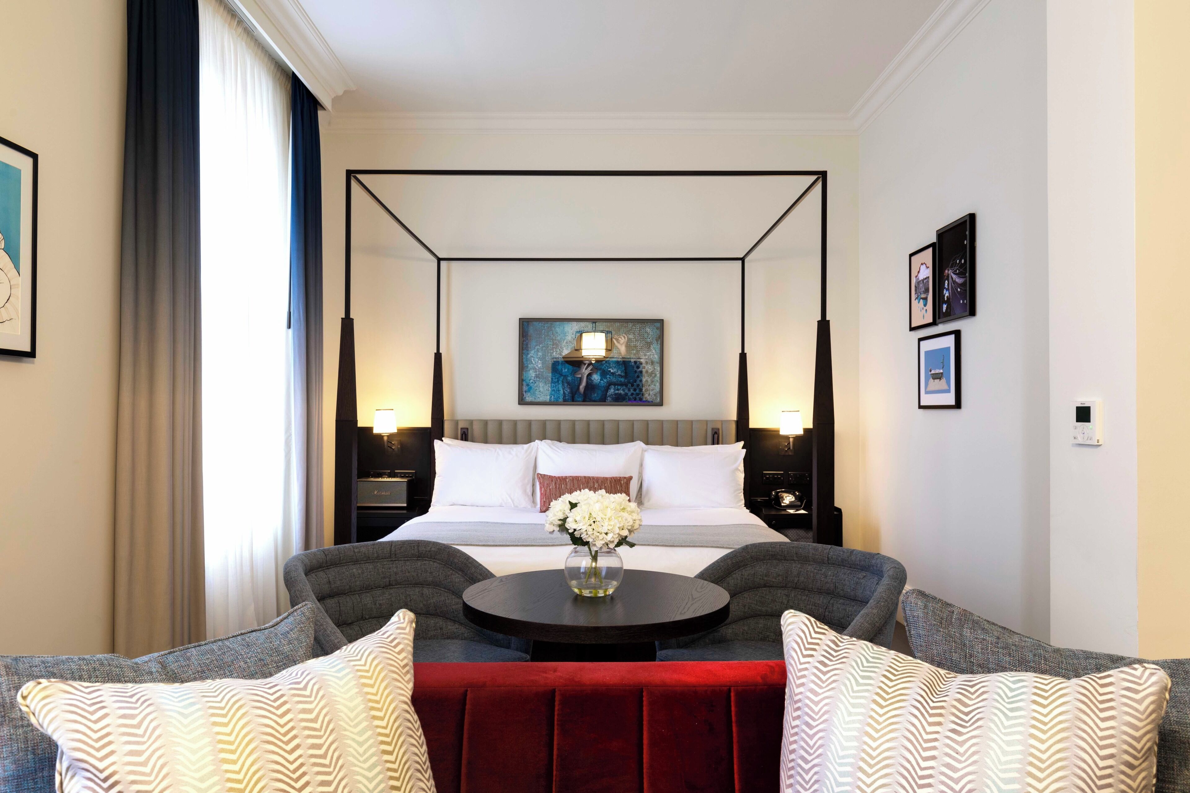 A four-poster bed with a vase of flowers, a round table, and two armchairs with a red ottoman.