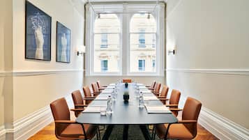 A conference room with a long table, brown chairs, and a view of buildings through large windows.