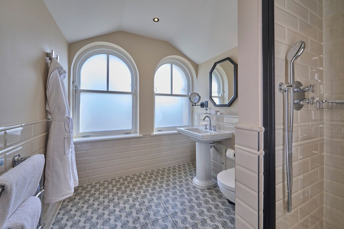 A bathroom with a bathtub, toilet, and sink, large windows, and patterned floor tiles.