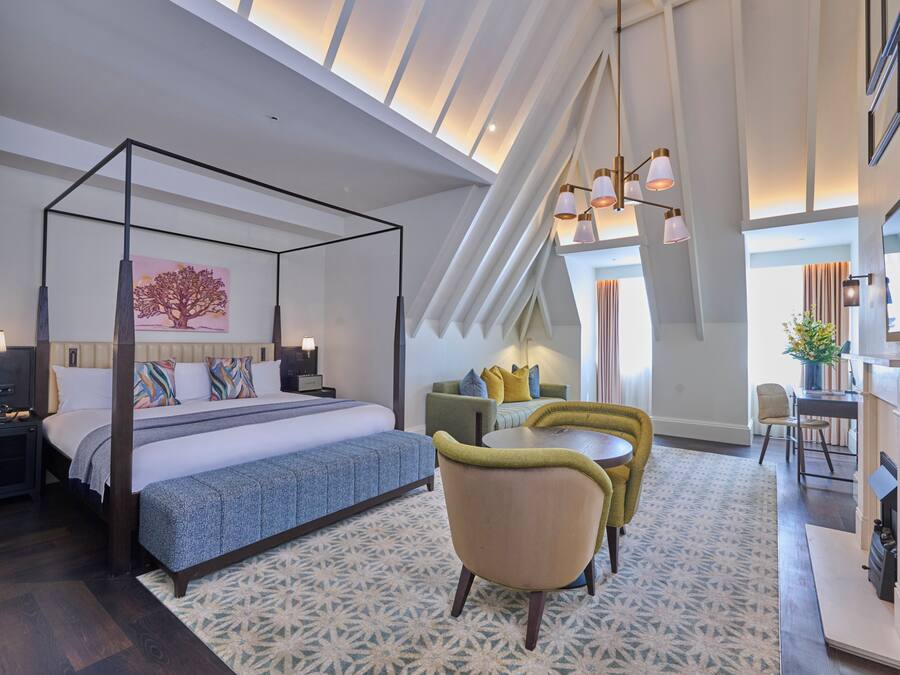 A modern hotel room with a four-poster bed, a dining area with a table and chairs, and a large window with a view.