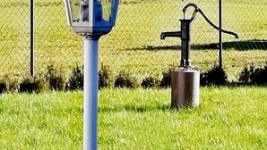Een blauwe straatlamp met een decoratieve bovenkant en een metalen voet, gelegen in een grasveld met een hek van gaas en bomen op de achtergrond.