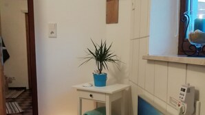 Een kleine kamer met een witte betegelde muur, een houten deur, een klein wit tafeltje met een plant in pot, een handdoek en een wandradiator.