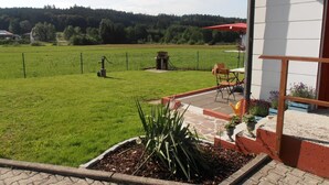 Een terras met een tafel en stoelen, een kleine tuin met planten, en een uitzicht op een groen veld en bomen in de verte.