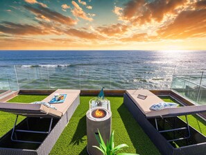 Una terraza en la azotea con dos sillones de descanso, un hogar para fuego y vista al océano al atardecer.