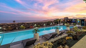 A poolside area with lounge chairs, a pool, and a view of the ocean at sunset.