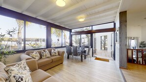 A spacious living area with a wooden ceiling, large windows offering a view of palm trees and a pool, a beige sofa with patterned cushions, and a dining area with chairs and a table.