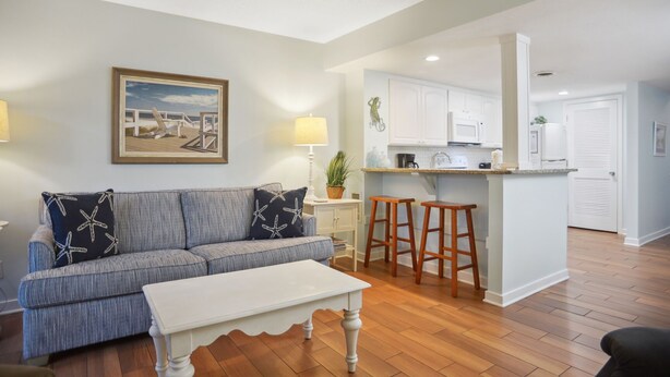 A living room with a sofa, a coffee table, and a kitchen area with a counter and stools. - Hilton Head Beach Club - 3 Bedroom w/ a Private Patio! (Hilton Head Island)