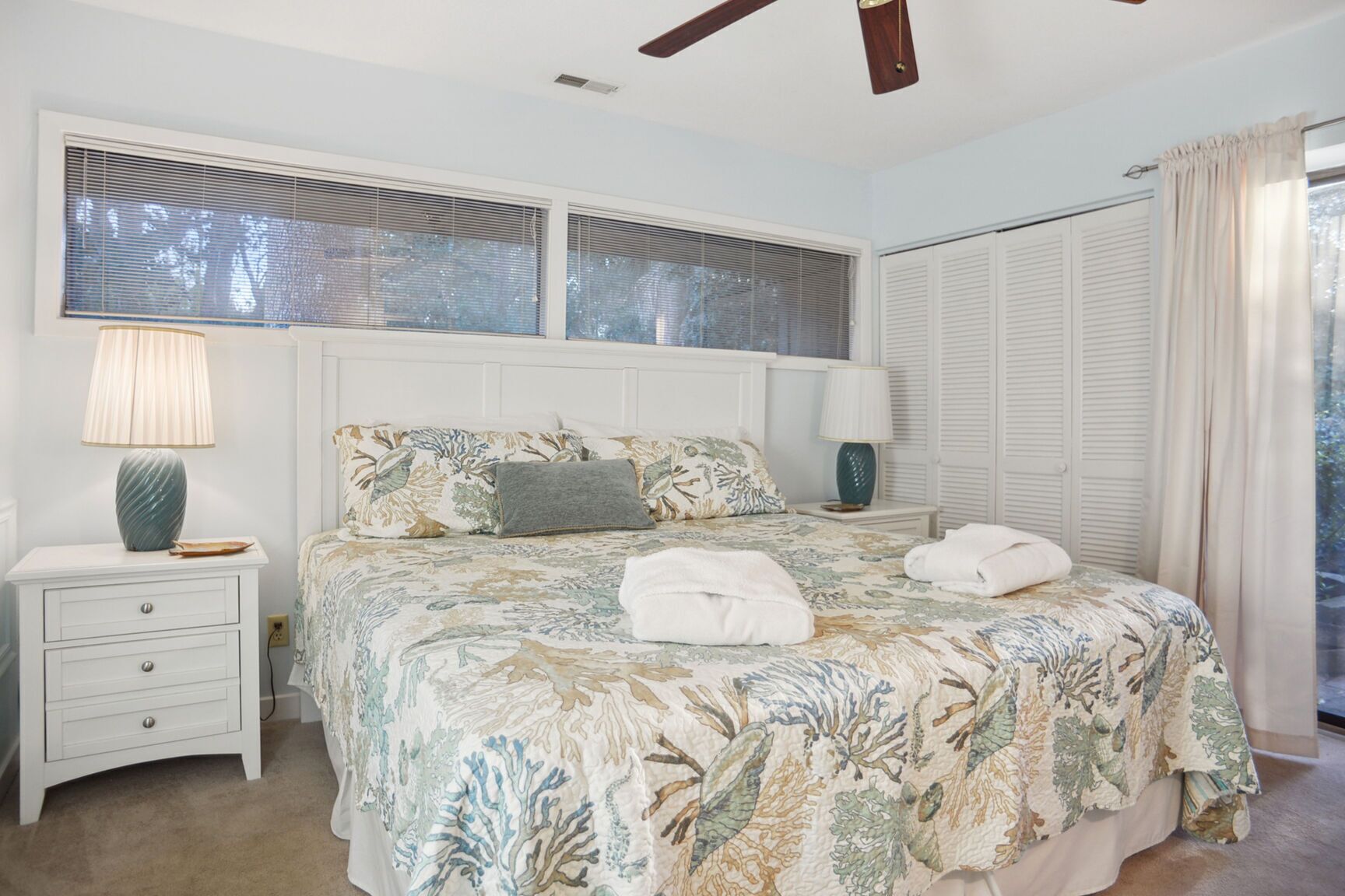 A bedroom with a bed, two bedside tables, a ceiling fan, a window with blinds, and a door with a curtain.