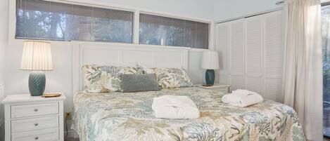 A bedroom with a bed, two bedside tables, a ceiling fan, a window with blinds, and a door with a curtain.