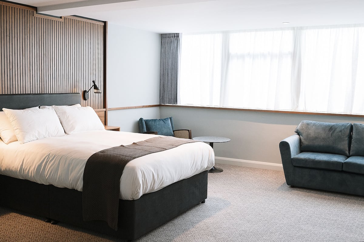 A modern hotel room with a large bed, a small round table, a sofa, and a window with curtains.
