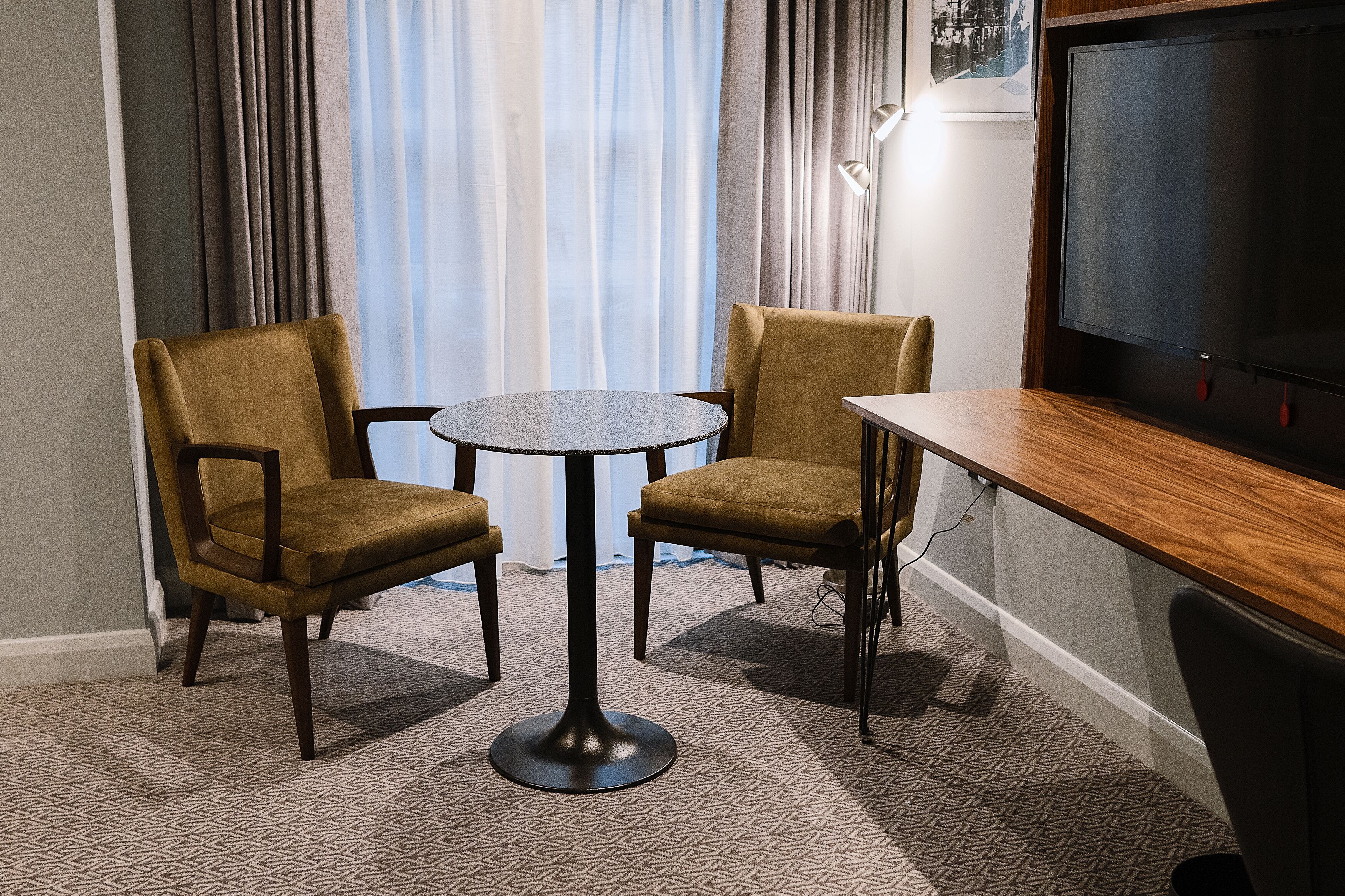 A room with two brown chairs, a small round table, a wooden desk, and a television on the wall.