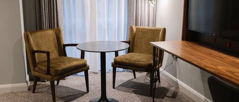 A room with two brown chairs, a small round table, a wooden desk, and a television on the wall.