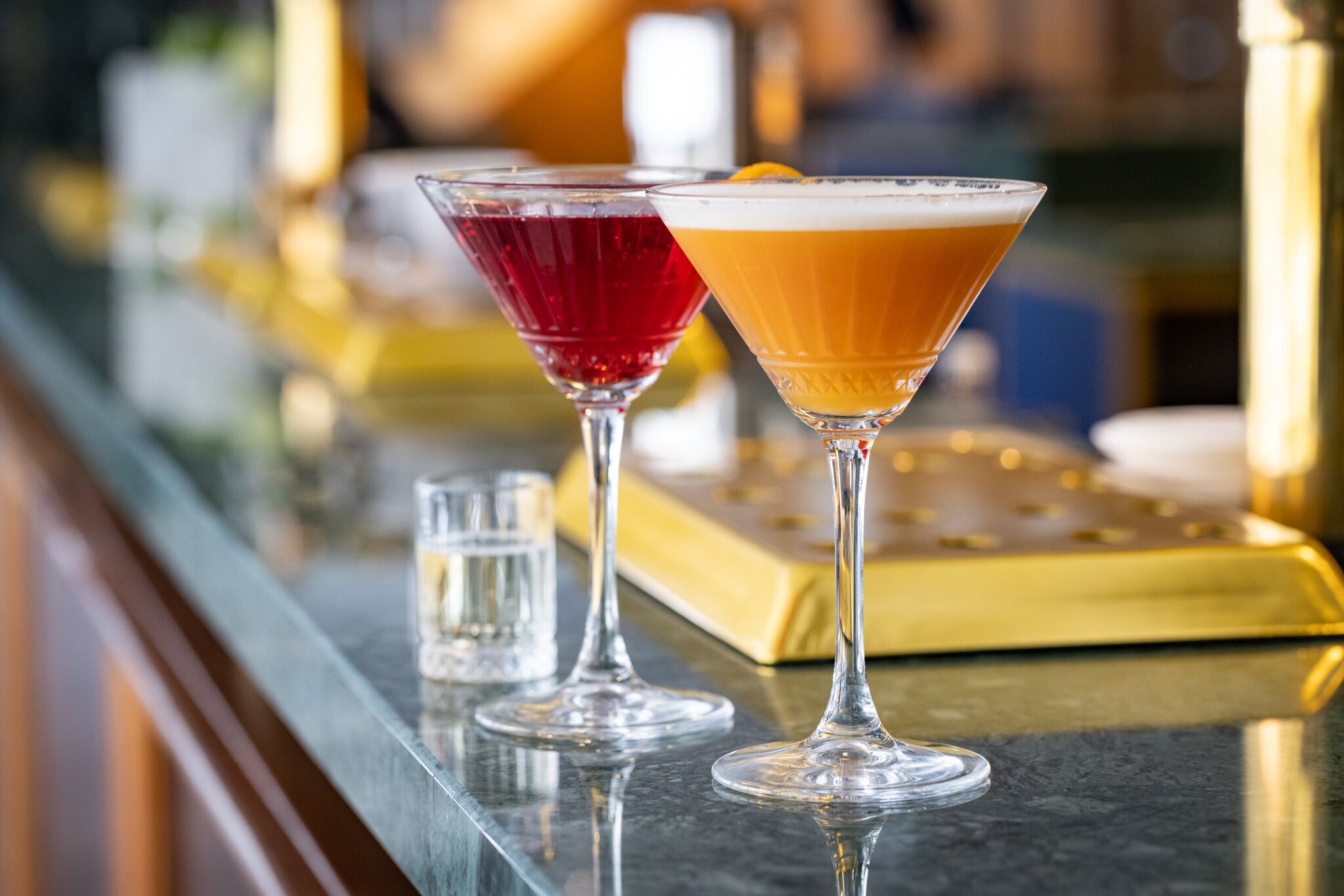Two martini glasses on a bar counter with a gold tray in the background.