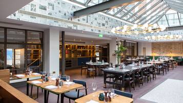 A spacious restaurant with multiple dining tables and chairs, a high ceiling with a skylight, and a brick wall in the background.