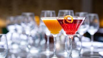 Two cocktails in martini glasses with an orange peel garnish, set on a bar with multiple glasses in the background.