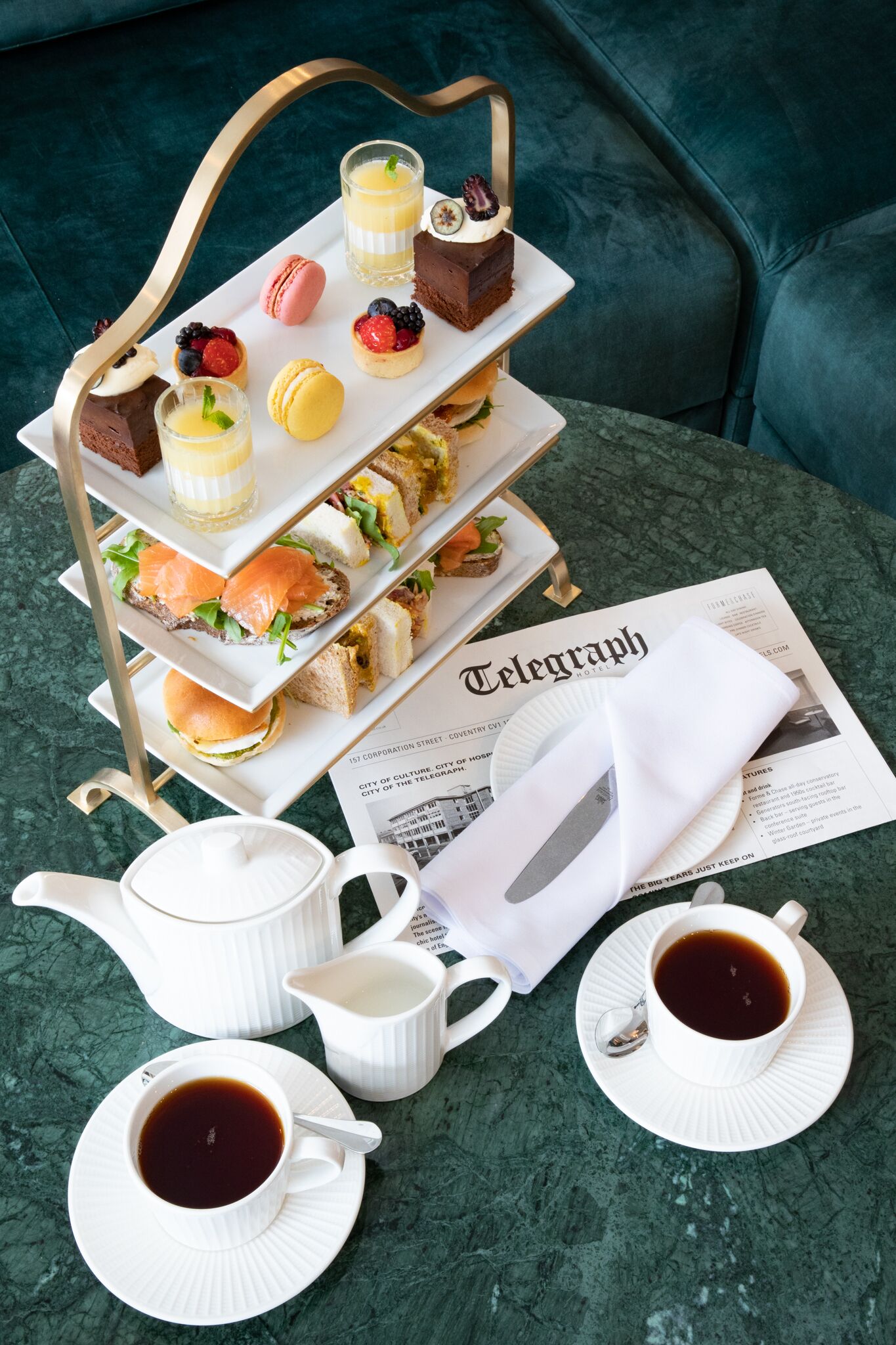 Un elegante plato escalonado con macarons, bayas y un sándwich, un periódico, una tetera y dos tazas de café sobre una mesa.