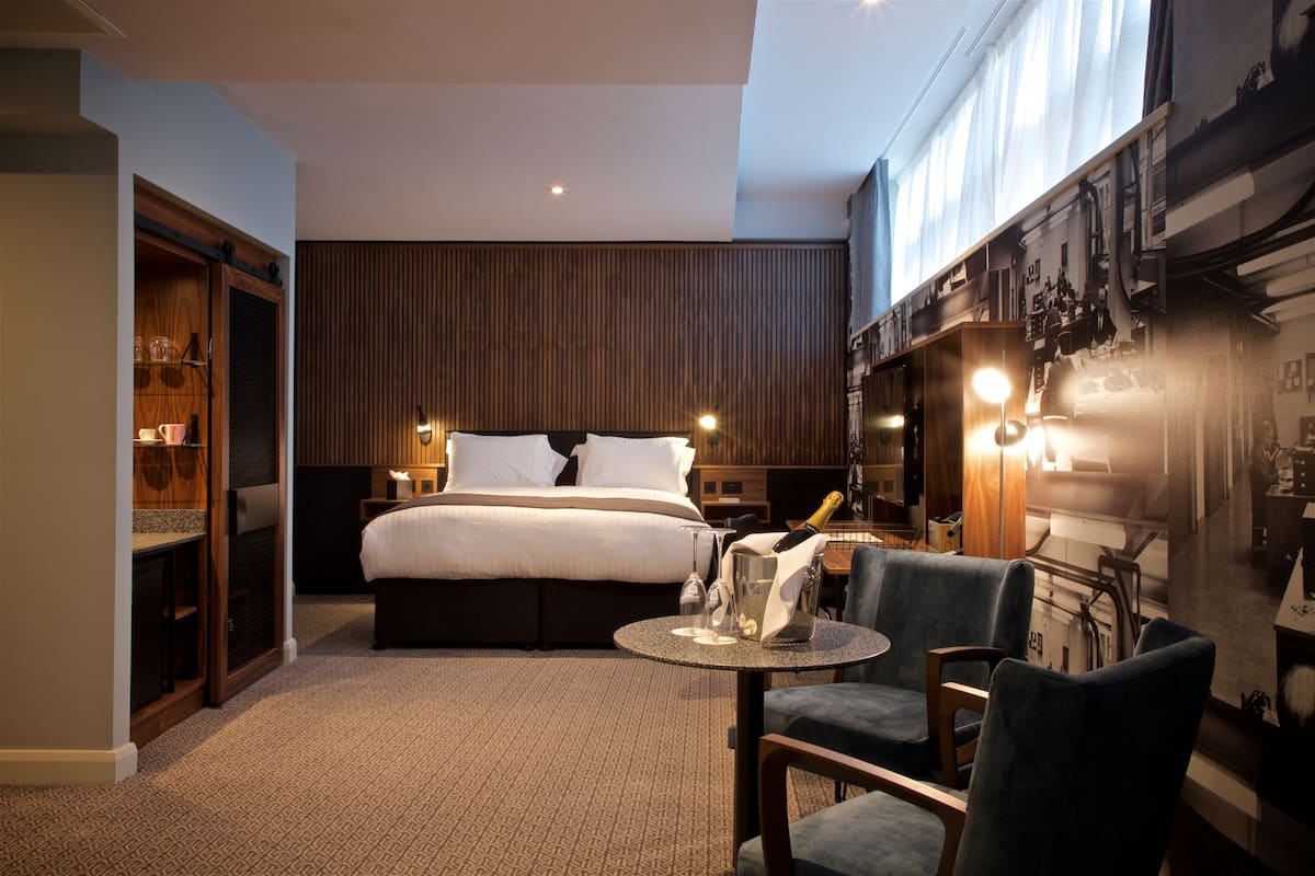 A hotel room with a large bed, a small round table with a bottle and glasses, a chair, and a minibar.